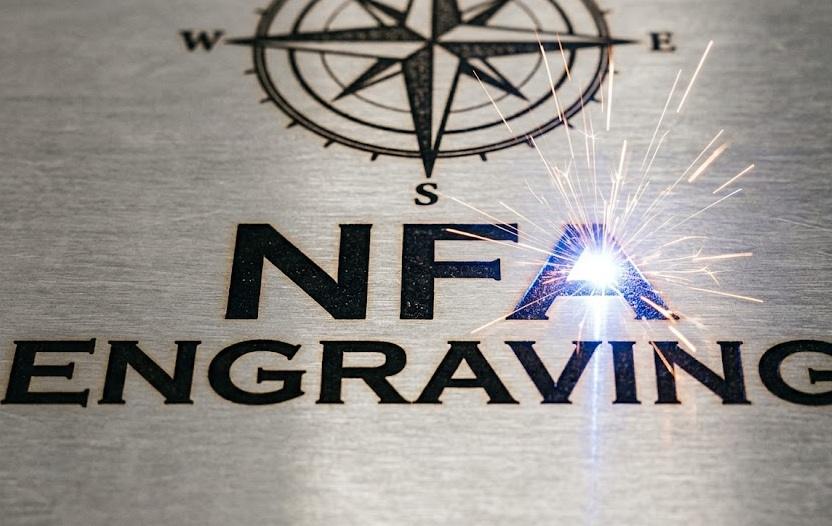 Close-up of metal engraving reading 'NFA ENGRAVING' with a compass rose logo above; a welding spark touches the letter A, highlighting the engraving process.