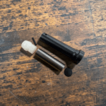 Disassembled small black plastic device with a metal sleeve, white plastic piece, tiny pin, and a round black cap on a wooden work surface. - Heavy Duty H2 4.6oz Steel Buffer With Reciprocating Internal Weight - Mid State Firearms , Mid State Firearms